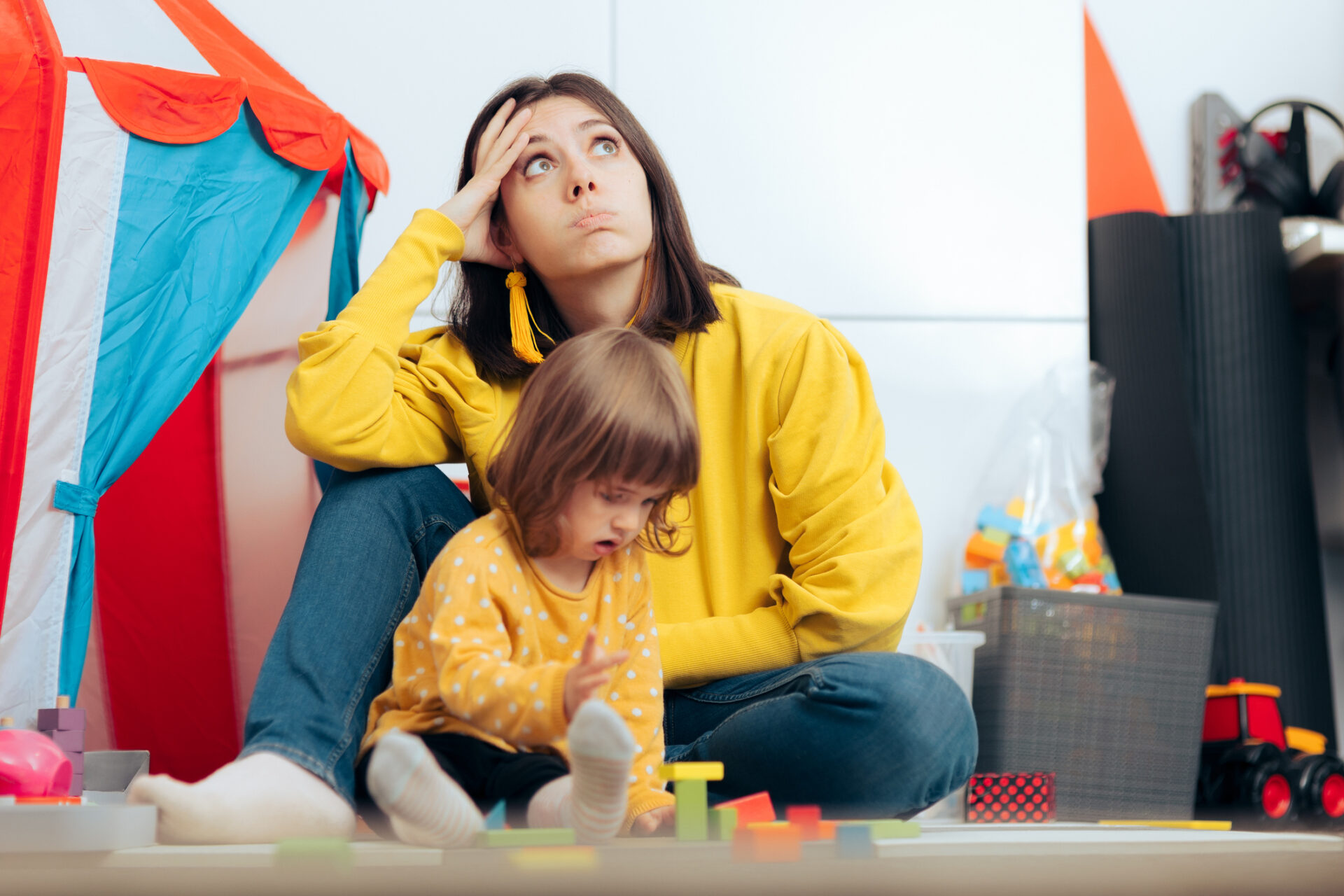 Mother in a yellow hoodie sits on the floor with a young child, looking worried in a cluttered playroom.